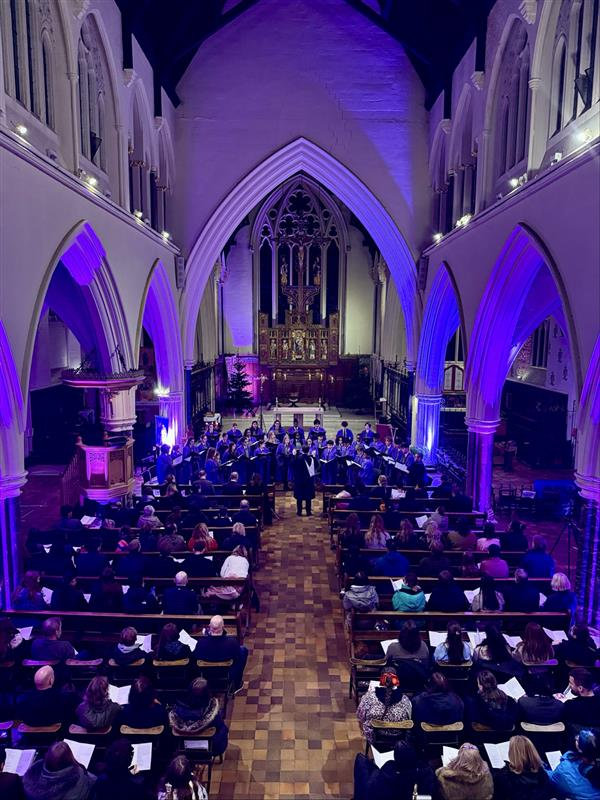 Cathedral interior with purple lighting, rows of seated people, and a choir or orchestra near the altar.