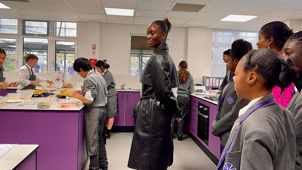 People standing in a purple school cafeteria, looking toward a counter with food trays.