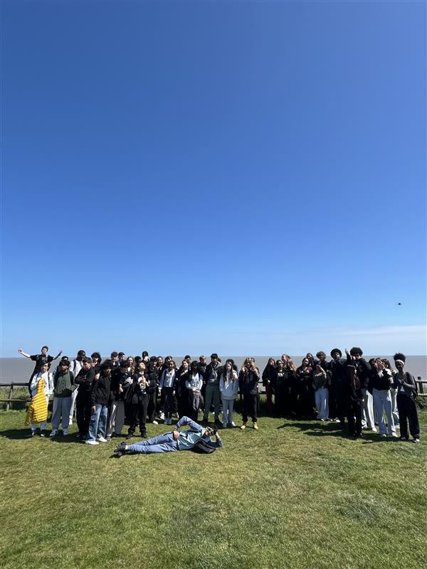 Large group standing on a grassy field under a clear blue sky, with one person lying in the foreground.