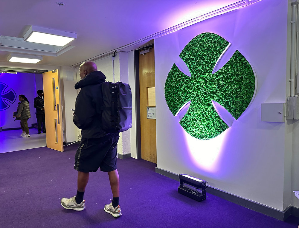 Hallway with purple carpet and wall lights; person in dark clothes stands near a glowing green wall logo.