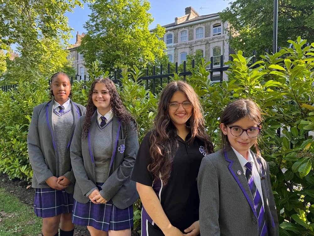 Four students in school uniforms standing outdoors in front of a building and greenery