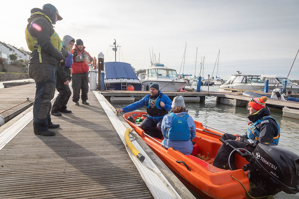 People in jackets on a dock. Three in an orange boat, others standing. Background of boats in a marina. Overcast sky, calm mood.