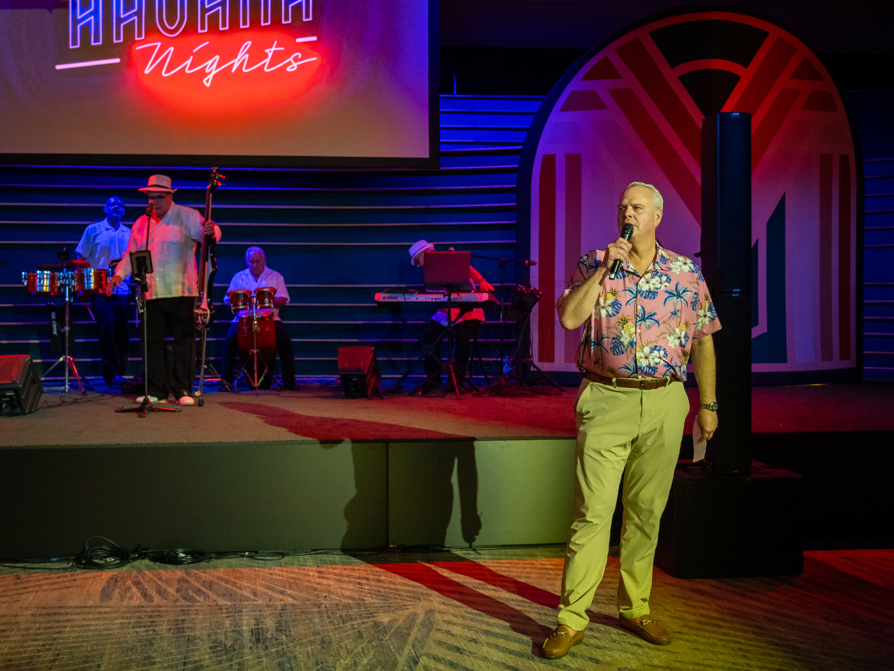 Man in a floral shirt sings into a microphone on a neon-lit stage with band; Havana Nights glows overhead.