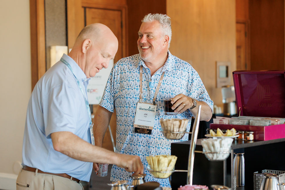 Two men chat at a hotel coffee bar; one smiles holding a drink, both wearing event badges, with The Invitational sign.