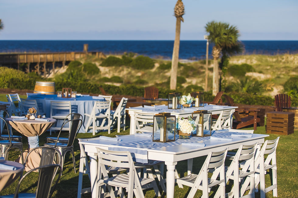 White tables with flowers and lanterns on a grassy area by the sea, under a blue sky. Chairs and palm trees in the background create a serene mood.