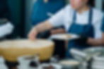 Chef in white coat and blue apron plates food from a giant Parmesan wheel at a buffet; focused kitchen scene.