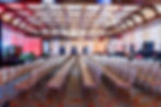 Empty ornate ballroom with rows of chairs facing a stage, chandeliers overhead, red curtains, and patterned carpet.