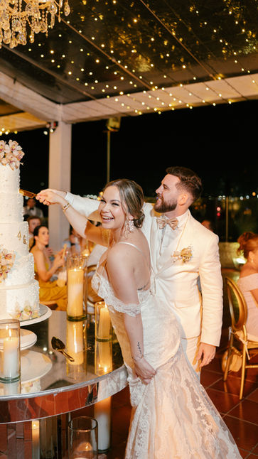 Novios sonrientes cortando pastel de bodas blanco con flores en recepción elegante.