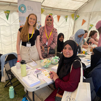 Women in a marquee with festive bunting in the background and an Elly Charity roller banner displaying the logo. Two women standing behind a table with papers in the foreground. A woman wearing a headscarf holds a cotton bag featuring the Elly Charity logo. Other women at the table engaging with members of the public