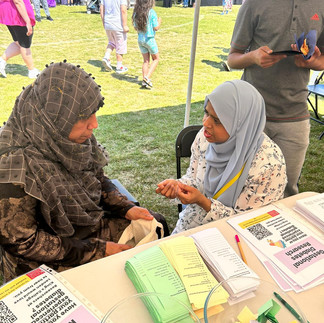Two women wearing headscarves engaged in a deep conversation. One is younger while the other is older. In the background, grass can be seen, indicating they are in a marquee. In the foreground, a table with papers is visible