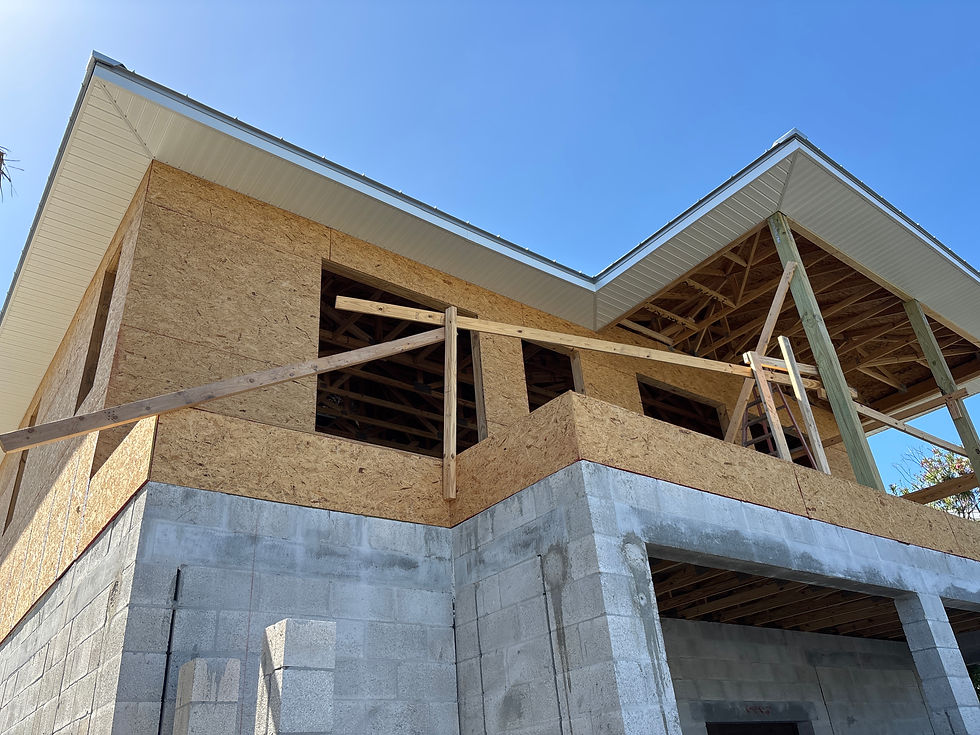 Unfinished two-story house under construction with exposed plywood, concrete block walls, and roof trusses against a clear blue sky