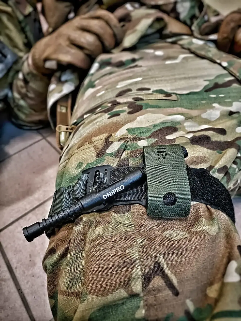 Un soldat en tenue camouflage allongé avec un garrot "DNIPRO" sur la jambe. Sol carrelé en arrière-plan, ambiance militaire.