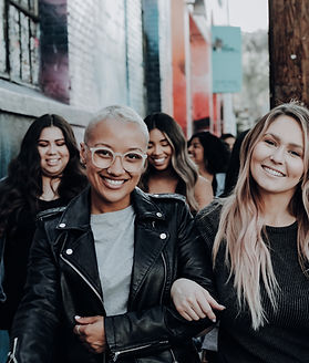 Several diverse women in black tops and jackets linking arms in a city, connecting throuhg an online Women's Support Group. Run by experienced therapists.