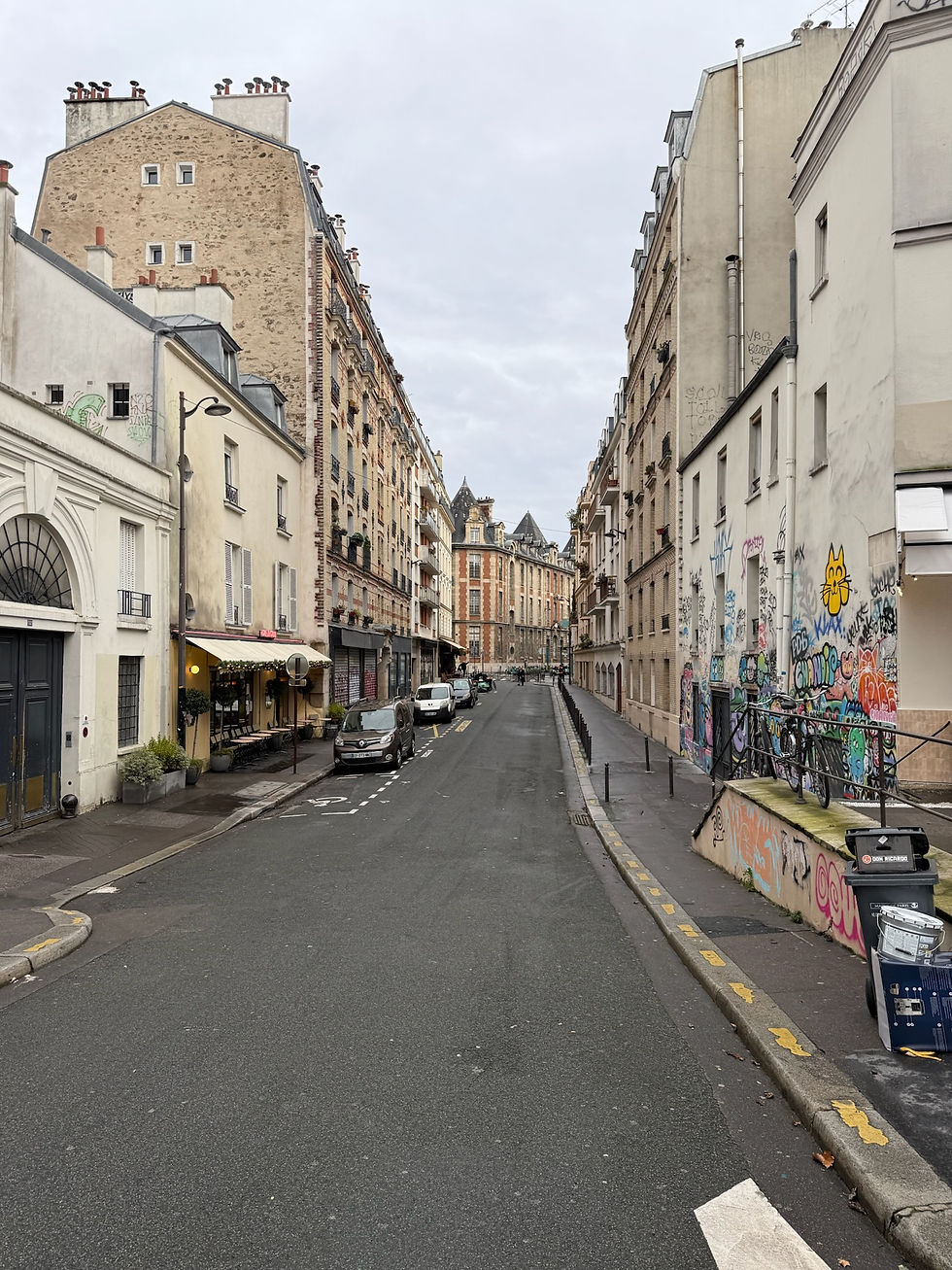 Narrow street lined with graffiti-covered buildings in an urban setting. Overcast sky, parked cars, and a distant person walking.