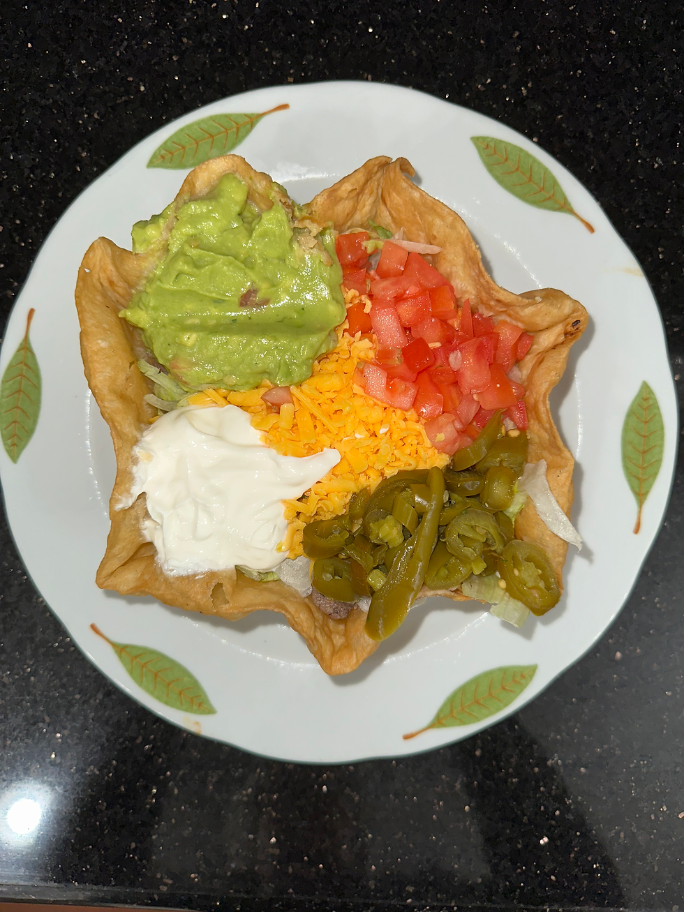 Taco salad in a crispy shell with guacamole, tomatoes, cheese, sour cream, and jalapeños on a leaf-patterned plate on a black countertop.
