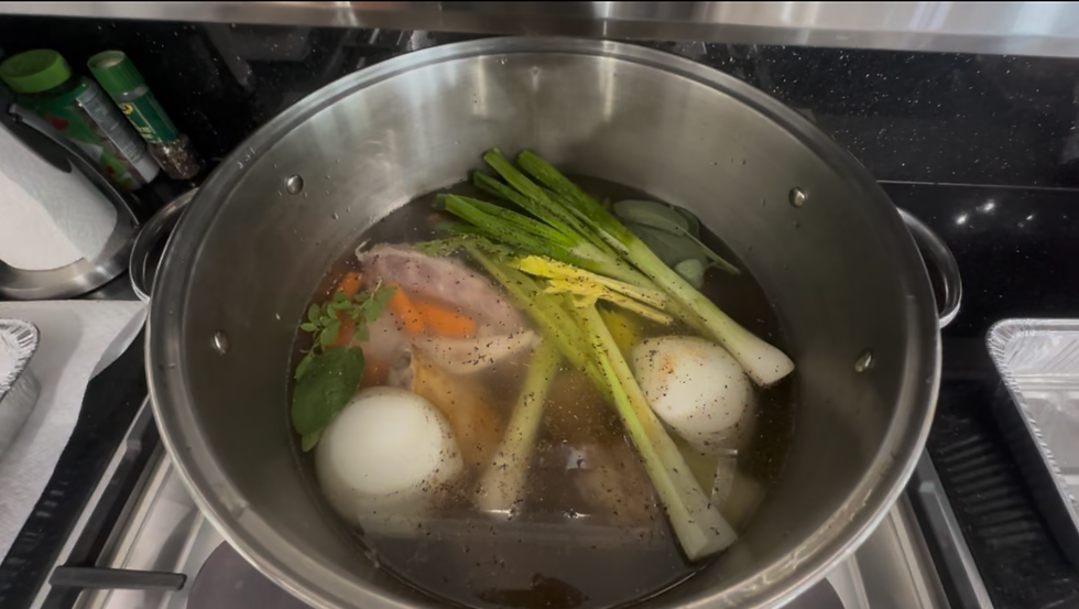 A pot filled with vegetables, herbs, and meat boiling on a stove. Visible ingredients include onions and green onions. Pepper is sprinkled on top.