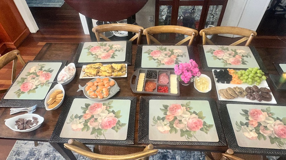 Dining table with floral placemats, assorted snacks, pink flowers, and plates of fruit, cheeses, crackers, cookies, and pastries. Cozy setting.