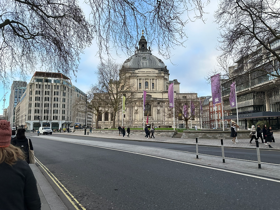 Methodist Central Hall in London stands at the center of the scene, its domed roof and classical stone façade framed by leafless winter trees and purple event banners. Pedestrians walk across the plaza and along the street in the foreground, with modern office buildings and traffic visible to the left under a pale blue, partly cloudy sky.