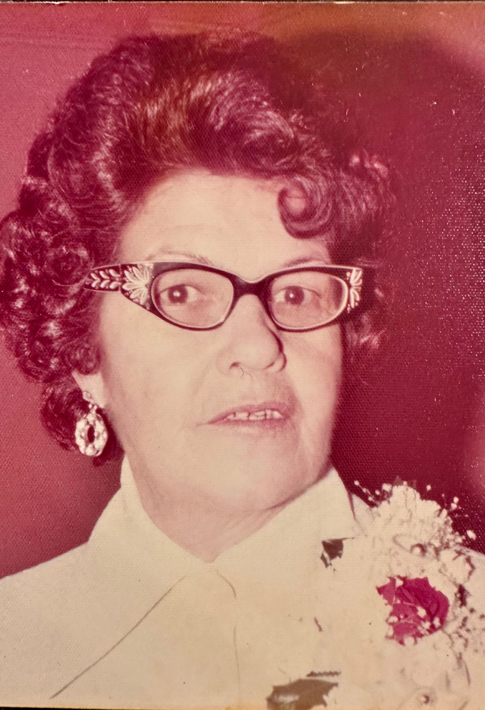 Woman with curly hair and glasses, wearing white, looks ahead. Red background, floral detail on dress, expressions are calm.