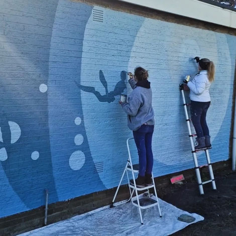 two women up ladders painting a blue underwater themed mural. 