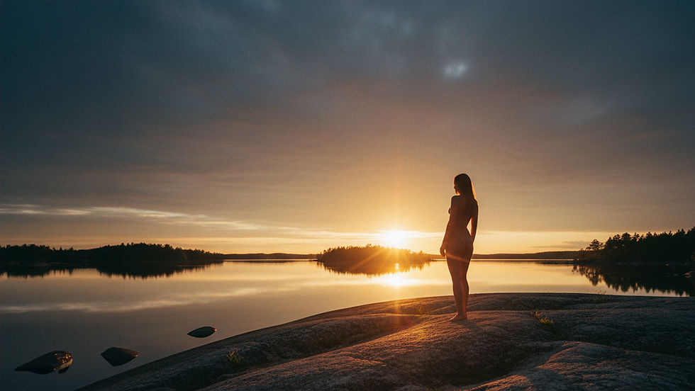 Taiteellinen alastonkuva siluettina Saimaan kalliorannalla auringonlaskun aikaan. Kultainen tunti heijastuu veden pinnasta ja valaisee mallin ääriviivat. Valokuvaaja Kuvaajankulma
