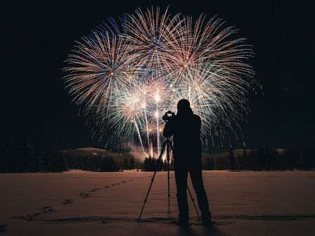Valokuvaaja kuvaamassa värikästä ilotulitusta järjestelmäkameralla ja jalustalla lumisessa talvimaisemassa.