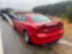 Red Dodge Charger on a wet roadside near a ramp, surrounded by grass and trees. Overcast sky and storage units in the background.