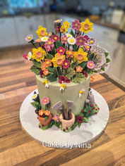 A vibrant birthday cake covered in lots and lots of sugar flowers, with gardening tools around the base