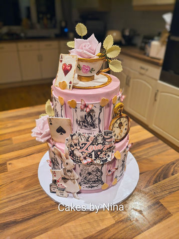 Beautiful two tier cake background is pink with lots of alice in wonderland reference the top has a teacup and saucer with sugar flowers