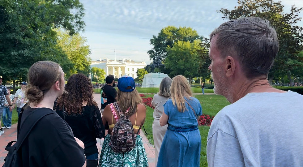 People stand in a park facing the White House. The scene is green with trees, red flowers, and a statue visible. Some wear casual clothes.