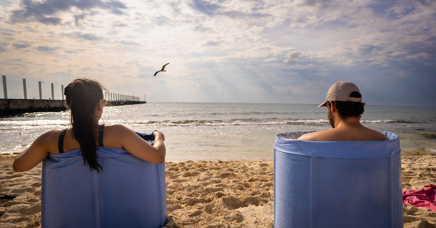 Rear view of male and female sitting in ice bath on beach, ocean background, sun rays beaming, bird flying by, Playa del Carmen, Mexico