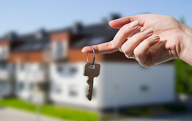 A house key dangles from an outstretched hand. A slightly out of focus house with white walls is in the background.