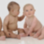 Two babies sitting on a white background; one touches the other's ear. Both wear nappies. The scene is innocent and playful, it shows a baby talking to a first born baby and the baby products it loves.