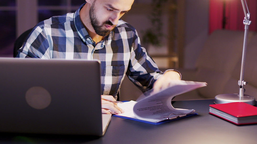 Man in plaid shirt focuses on papers, working at desk with laptop and lamp. Evening setting, warm lighting, casual and studious mood.