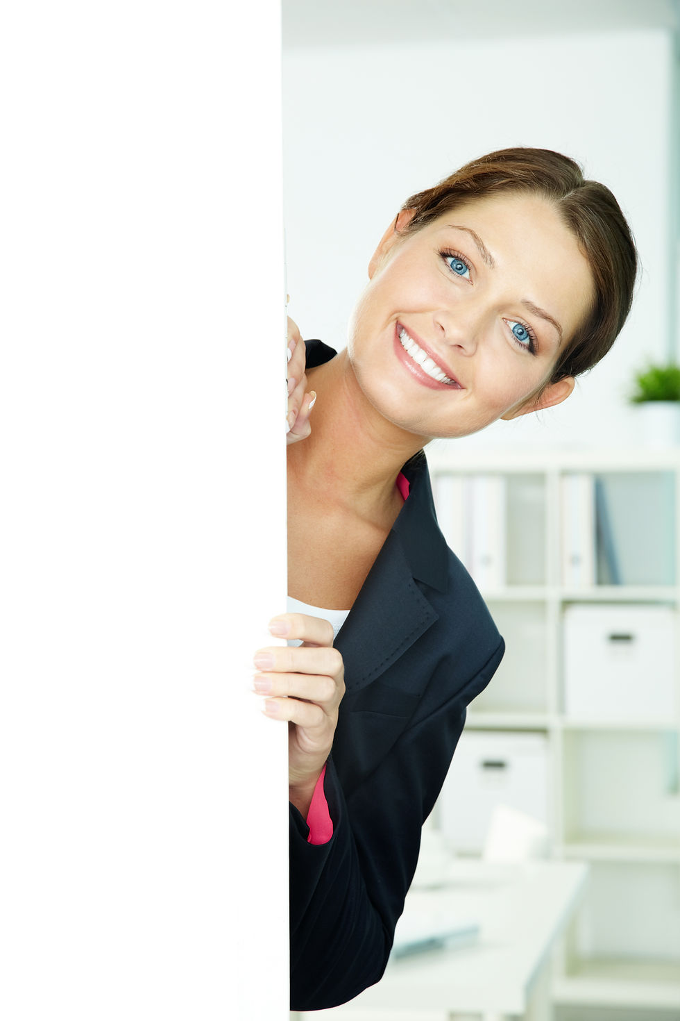 Smiling woman in a suit peeks from behind a white panel in a bright office setting with shelves and plants. Cheerful and welcoming mood.