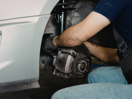 Mechanic in jeans and gloves fixes a car's brake system, with focus on the wheel area. The setting suggests a garage environment.