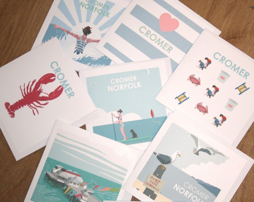 selection of greetings cards with designs relating to Cromer. Red lobster, crabs, Cromer Pier, seagulls, woman and dog on paddleboard, woman in striped bathing suit in the sea