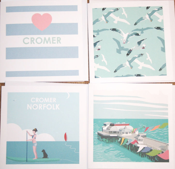 selection of greetings cards with designs relating to Cromer. Crabs, Cromer Pier, seagulls, woman and dog on paddleboad.