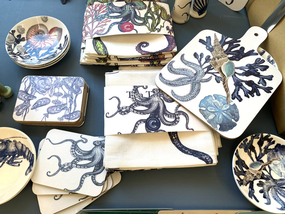a selection of teatowels, cutting board, small dishes and coasters all white and printed with navy, purple and green realistic images of octopus, shells, coral and starfish