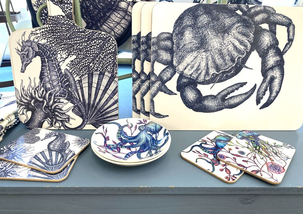 white coasters with navy blue realistic crab printed alongside coasters and small dishes with realistic octopus print, and a cutting board with navy blue realistic seahorse and scallop shell design