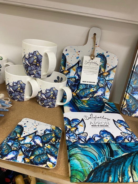 white china mugs, coasters and an apron printed with realistic dark blue/green mussel shells with Dollyhotdogs branding