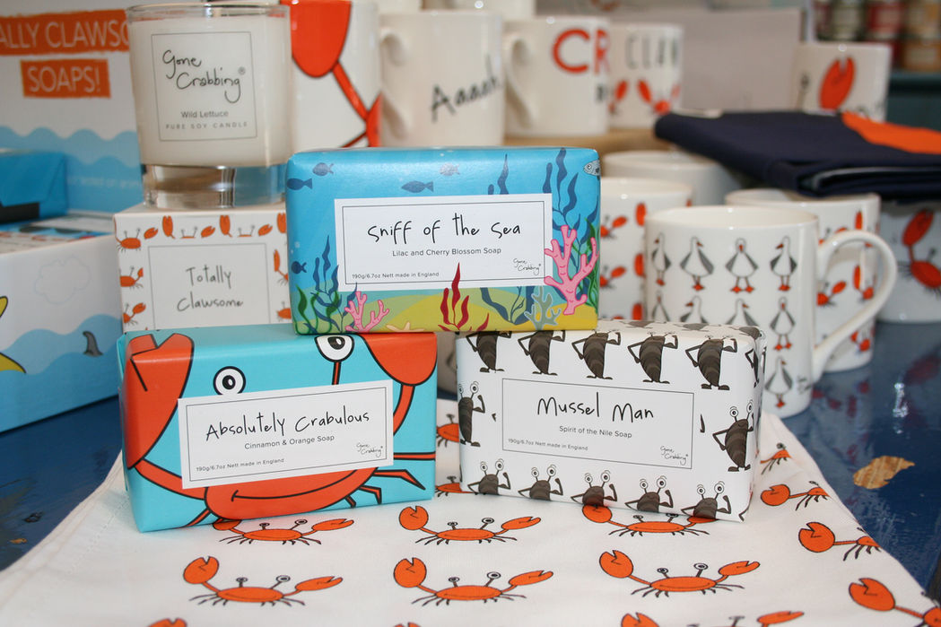 white teatowel with friendly crab logo topped with three different bars of soap, one showing a cheeful crab, one showing an undersea scene with coral and the third with mussels on with Gone Crabbing branding