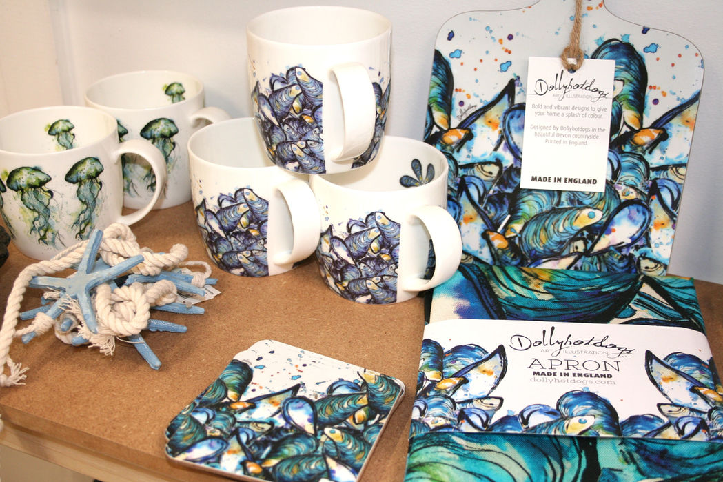 white china mugs, coasters and an apron printed with realistic dark blue/green mussel shells alongside additional white china mugs with green jellyfish design with Dollyhotdogs branding