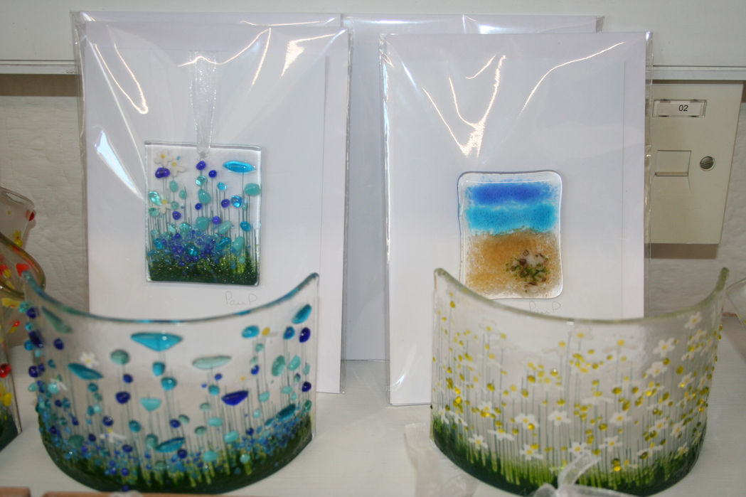 two white greetings cards with a small glass rectangle one decorated with a meadow scene with blue flowers and the other with the blue sky and golden sand of a beach. these are upright behind two curved glass candle holders one with blue flowers and the other with white and yellow flowers