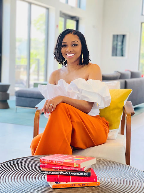 Picture of Dr. Samantha Rae sitting down with books in front of her. She is smiling and has on a white top with orange pants