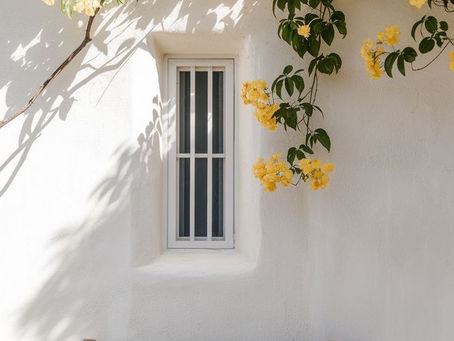 Parede branca, com uma unica janela, embaixo um banco de madeira e uma arvore com flores amarela acima da janela