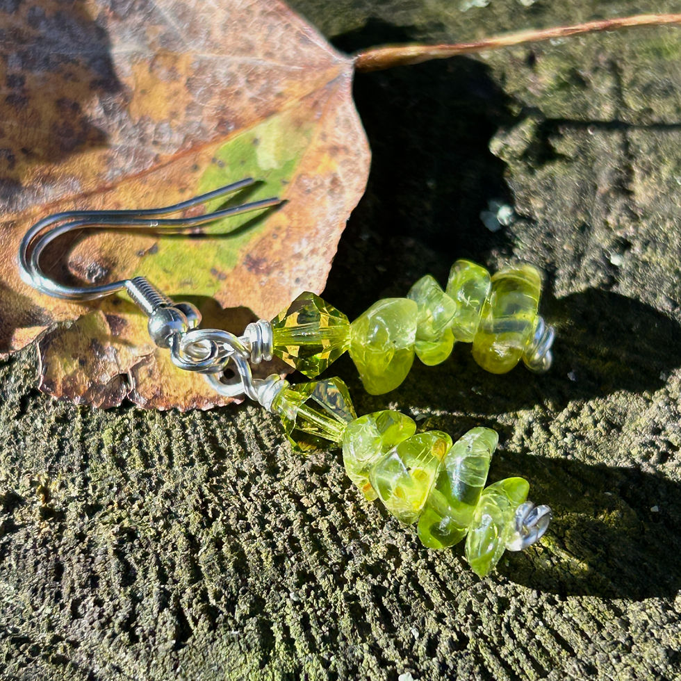 Thumbnail: Green chip natural form peridot earrings made with stainless steel on a wood stump with fall leaves