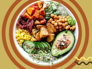 An overhead view of a vibrant vegan bowl featuring a variety of ingredients including diced roasted potatoes, chickpeas, sliced cucumbers, corn, sundried tomatoes, sprouts, and a halved avocado topped with a creamy sauce, all set against a background of concentric tan and brown circles.