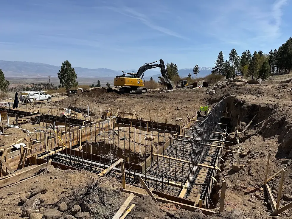 Construction site in a rural area with metal rebar and trenches visible. Excavator and truck in the background. Clear blue sky.