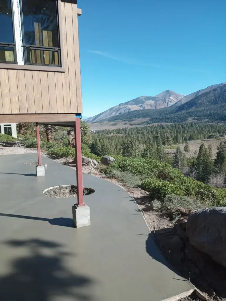 A wooden house on stilts overlooks a scenic mountain landscape. Fresh concrete path surrounds the house. Clear blue sky in the background.